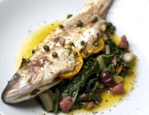 Branzino Mediterranean Sea Bass - Whole, Gutted, Scaled cooked and plated with leafy greens and capers.